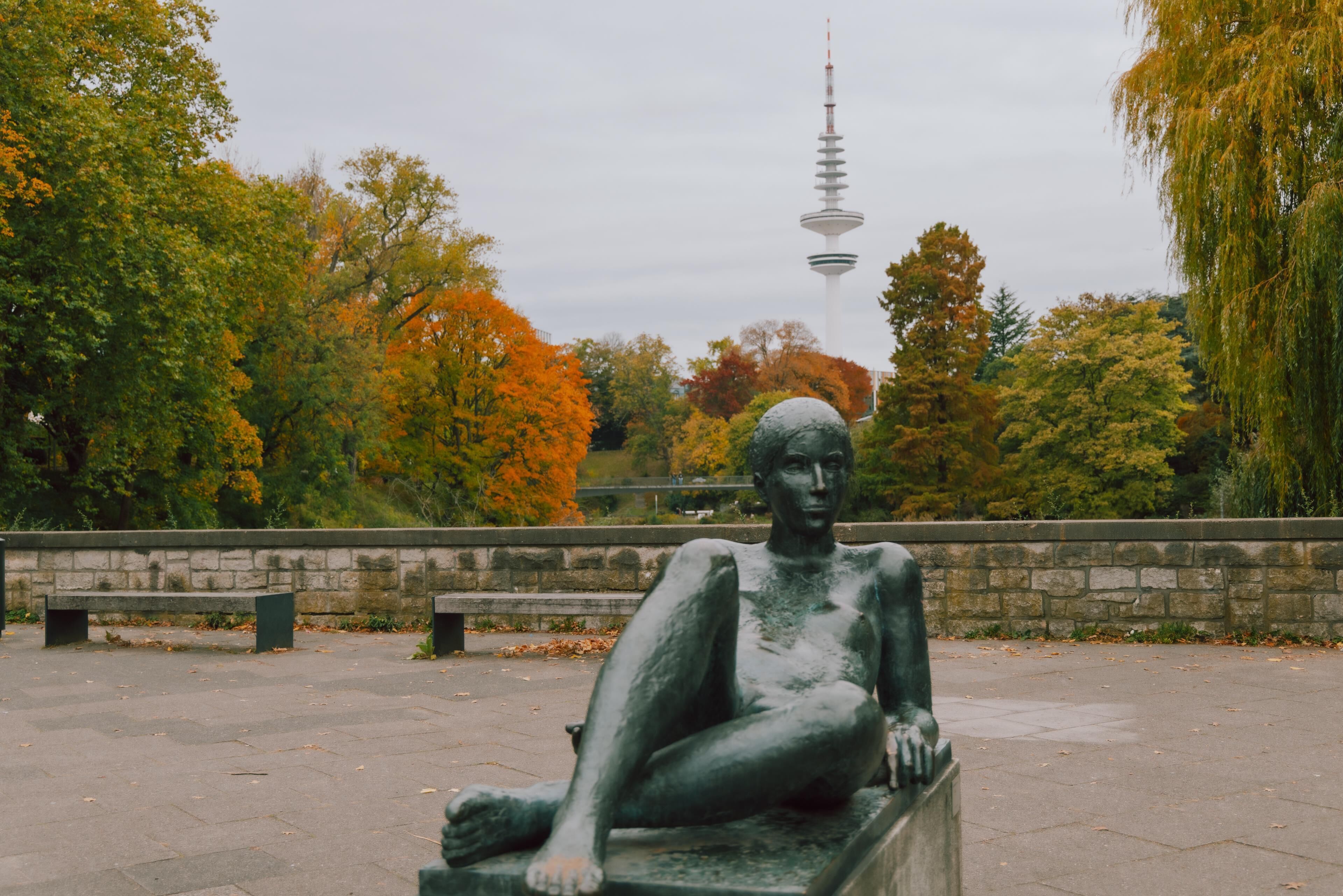 Eine Statue einer sitzenden Person in einem Park mit Bäumen und einem Turm im Hintergrund.
