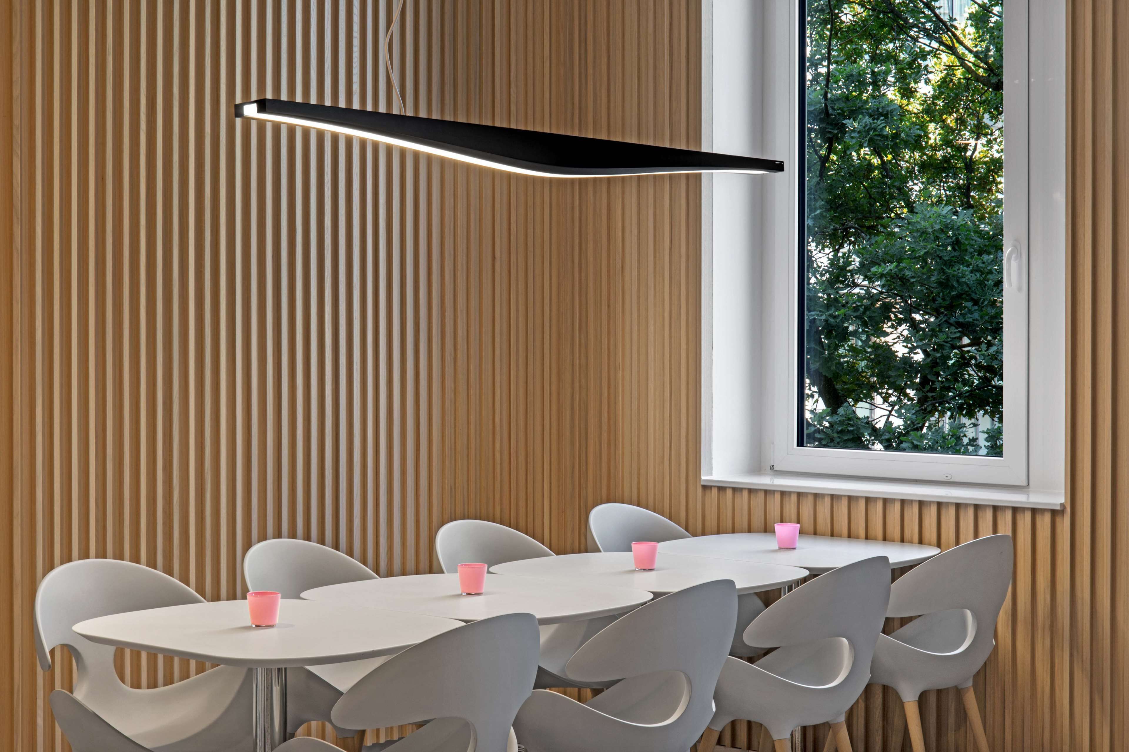 A modern dining area with a round table and white chairs, a wooden paneled wall, and a window with a view of trees.