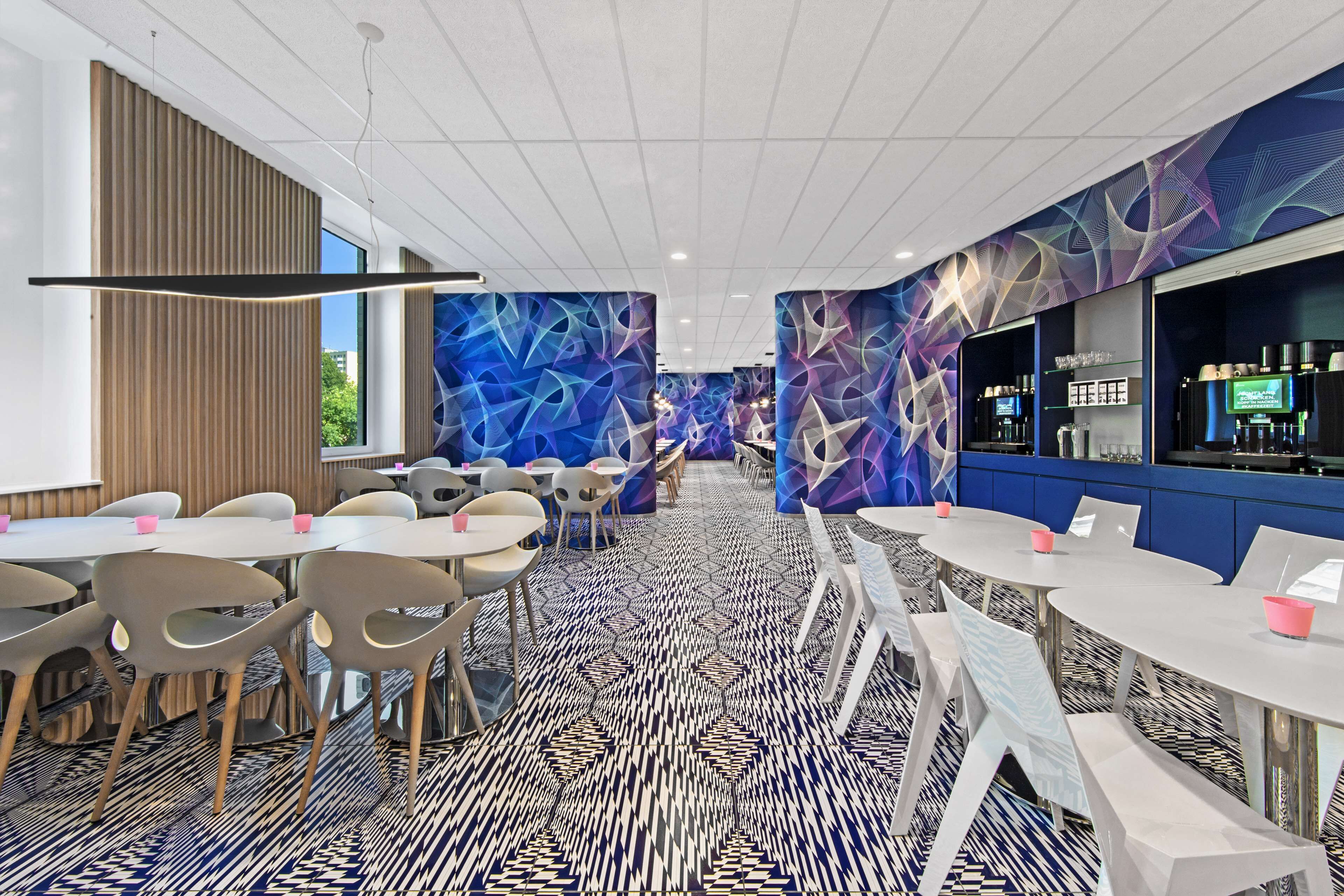 Eine moderne Hotelhalle mit runden Tischen und Stühlen, einem gemusterten Teppich und einer Bar mit einem digitalen Bildschirm.