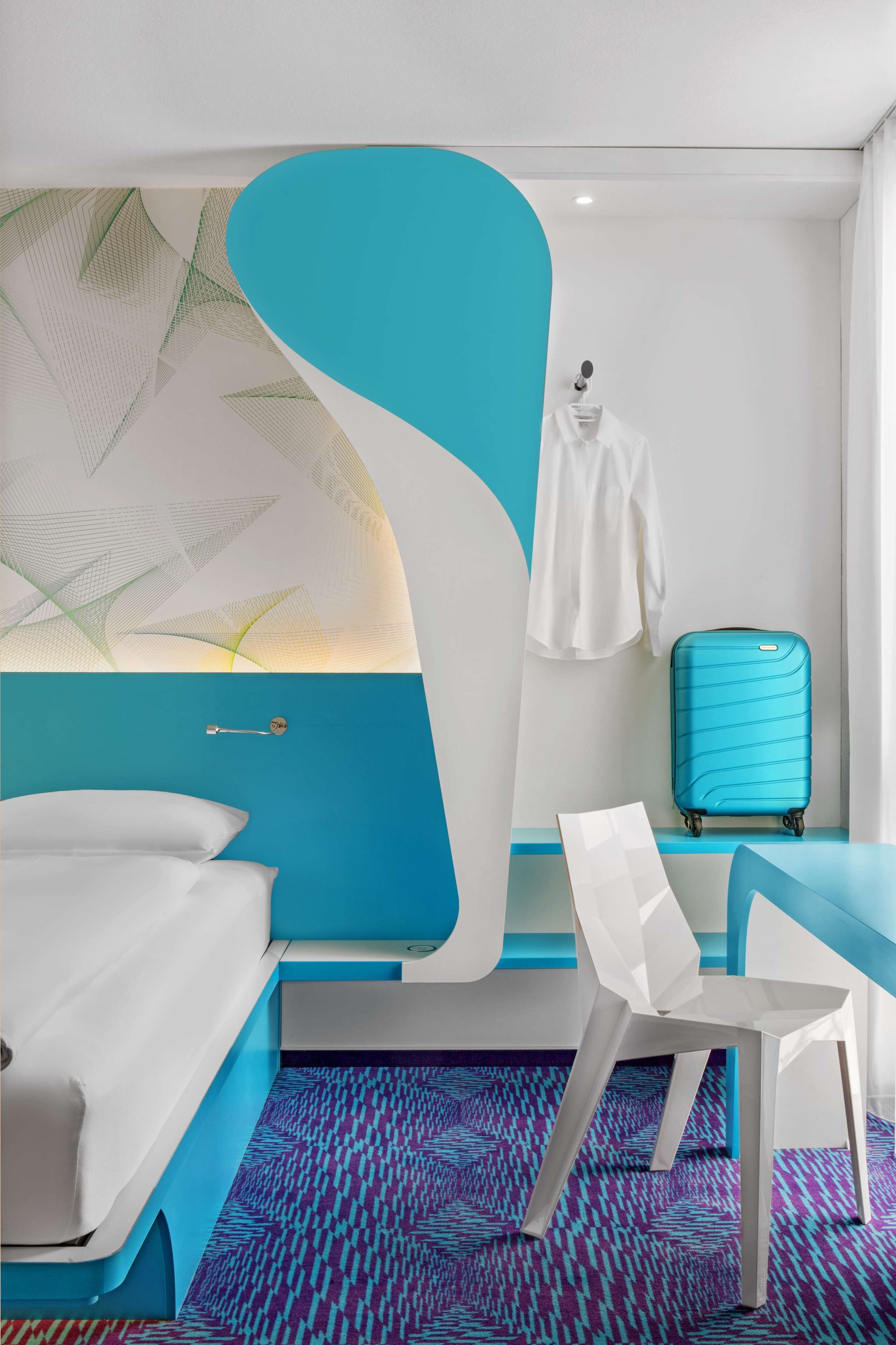 Camera d'albergo moderna con arredamento nei toni del blu e del bianco, un letto, una scrivania, una sedia, una valigia e un cappotto appeso.