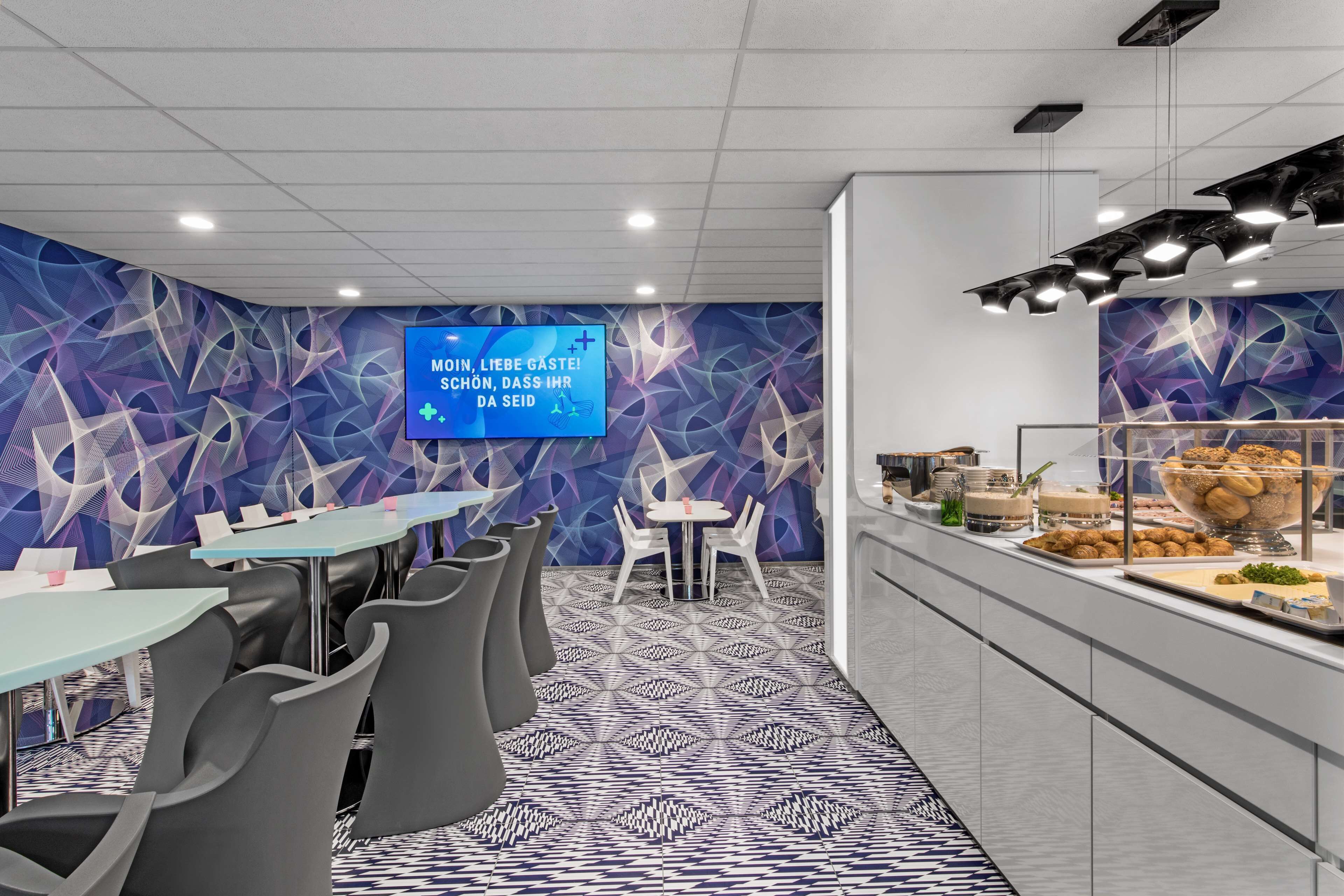 A modern dining area with a geometric patterned wall, a digital screen displaying a message, and a buffet counter with various food items.