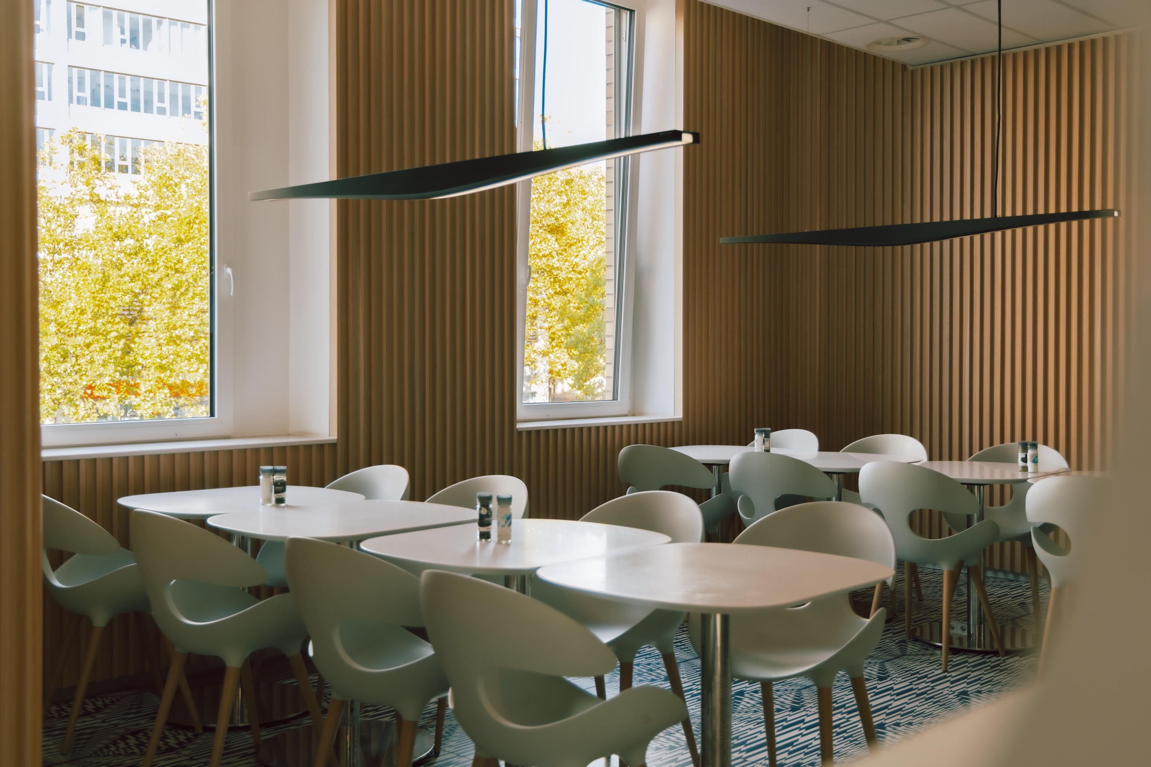 Un área de comedor moderna con mesas redondas y sillas de colores claros, ventanas amplias con persianas verticales y una alfombra estampada.