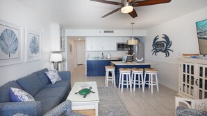 A modern living room with a grey sofa, a white coffee table, and a blue patterned rug. There is a kitchen area with blue cabinets and a white countertop. - COUPLES ROMANTIC GETAWAY, OCEANFRONT, HIGH-END DECOR, ADJUST. KING, FREE PARKING (Myrtle Beach)