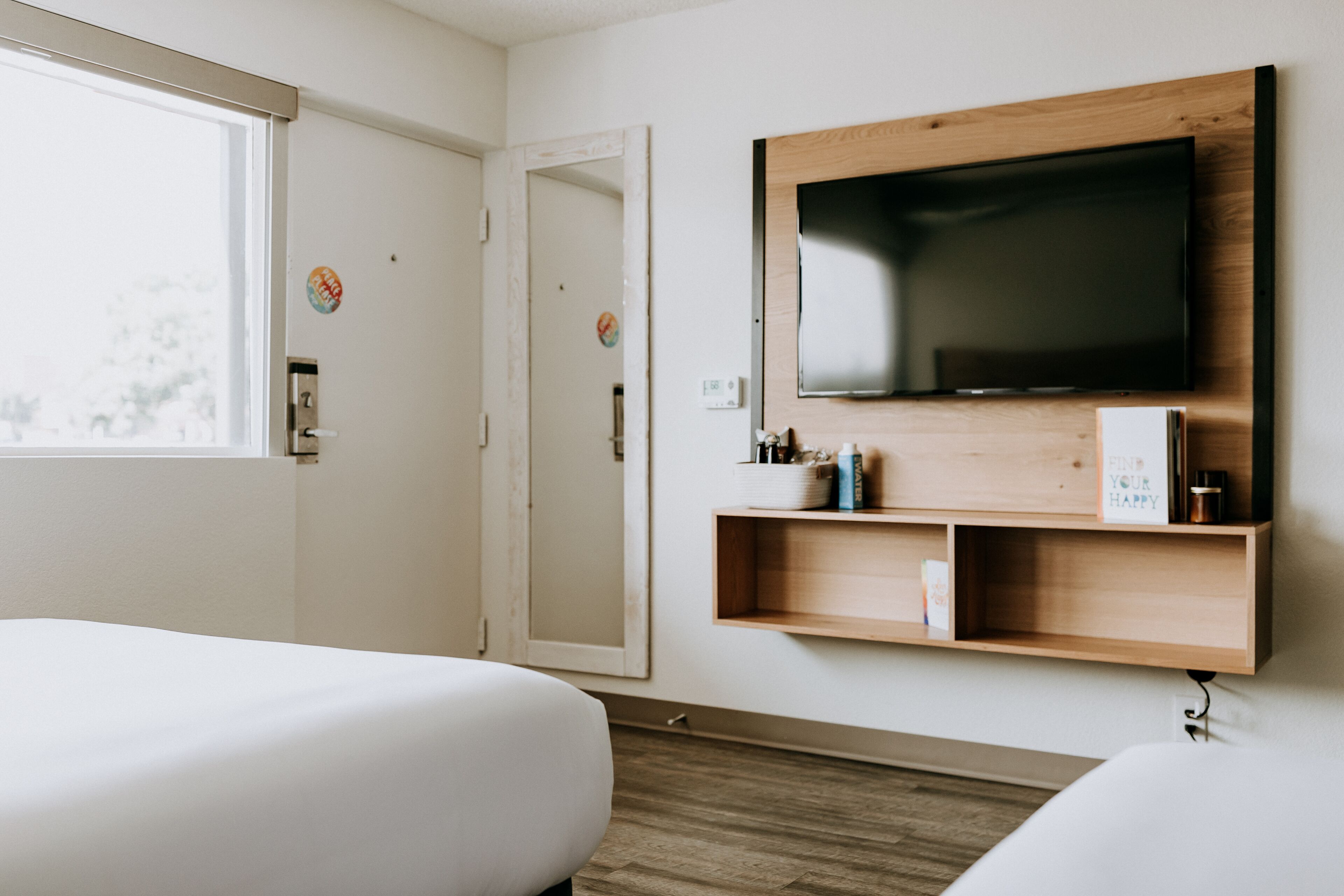 A hotel room with two beds, a TV mounted on the wall, a small shelf with items, and a window.