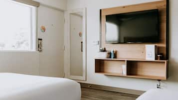 A hotel room with two beds, a TV mounted on the wall, a small shelf with items, and a window.