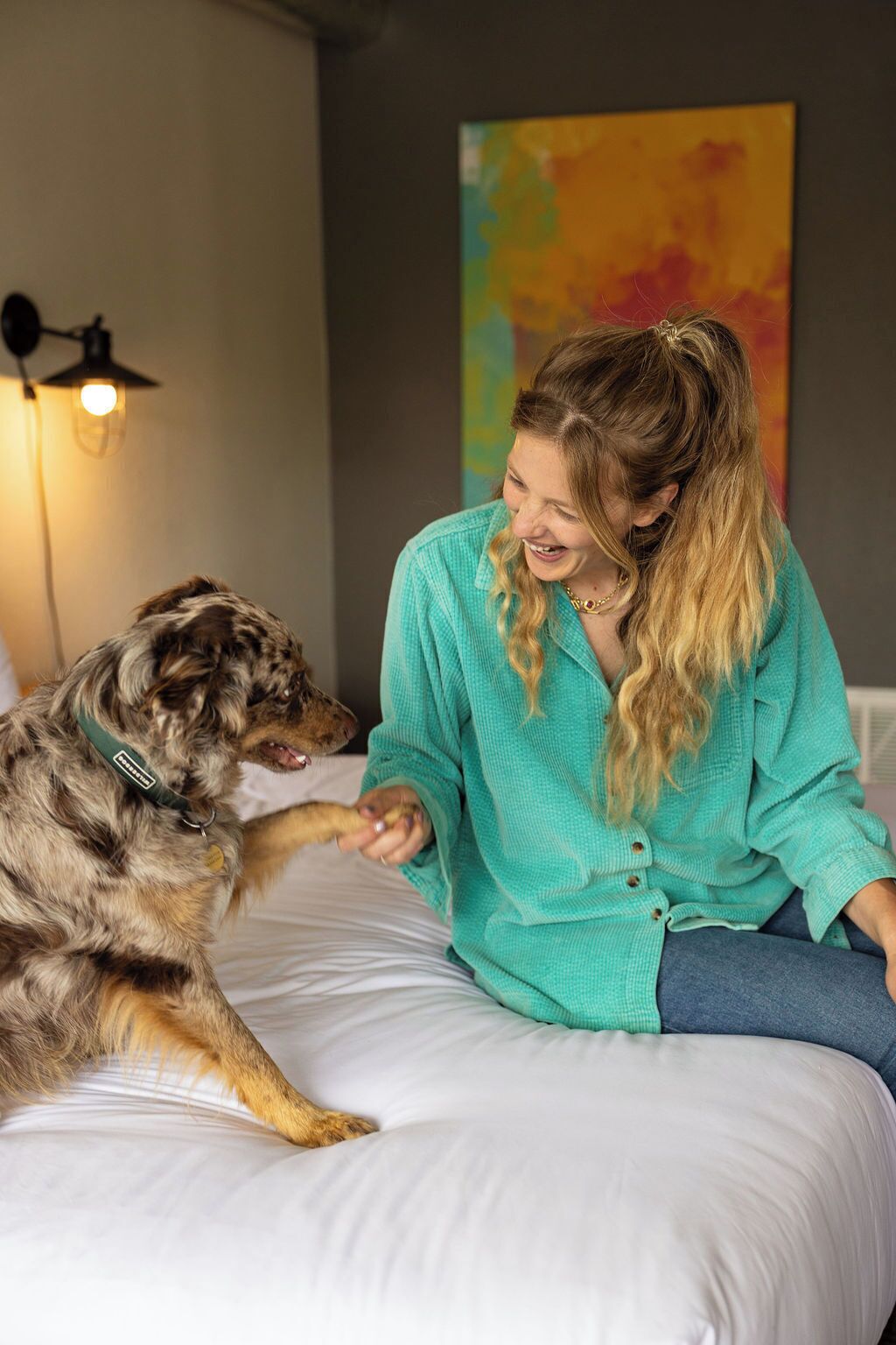 A person sitting on a bed with a dog, a lamp, and a colorful abstract painting in the background.