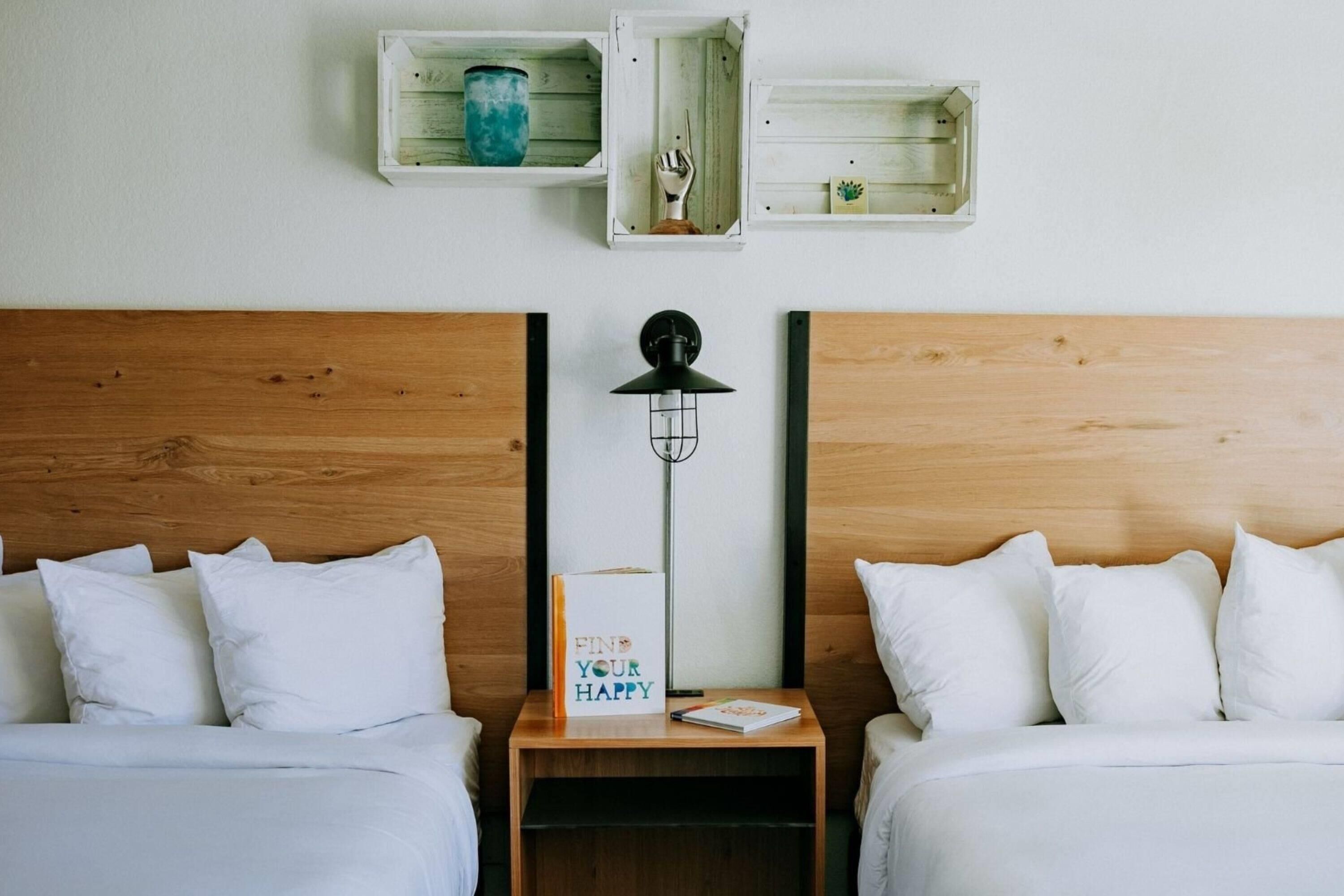 Habitación de hotel con dos camas, una mesita de noche, una lámpara de pared y dos cajas de almacenamiento abiertas con objetos decorativos.