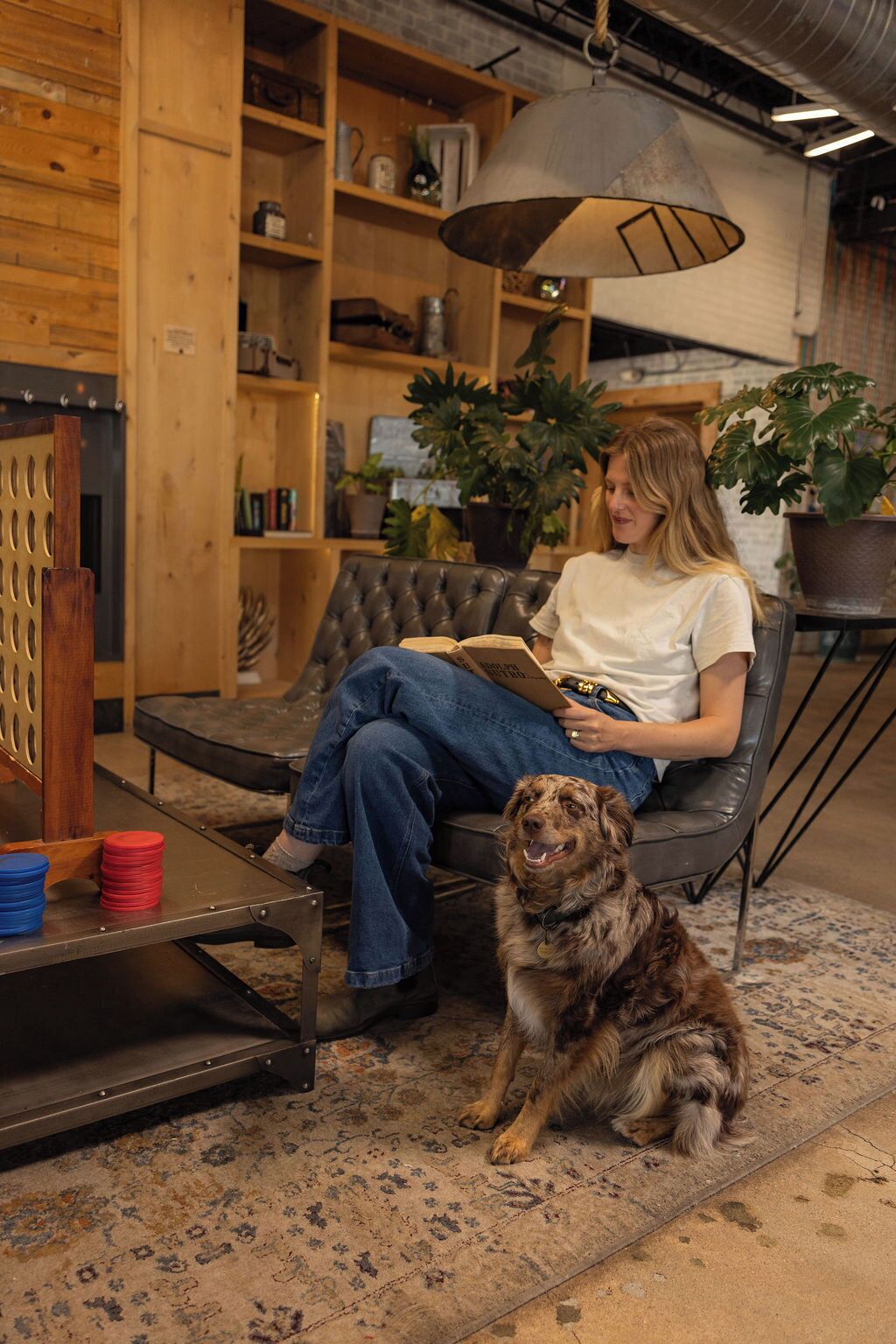A person sitting on a leather couch reading a book, with a dog beside them.
