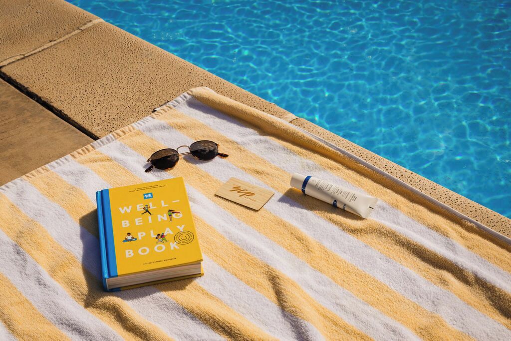 A poolside setting with a towel, a book titled 'Well-Being Play Book,' sunglasses, and a bottle.