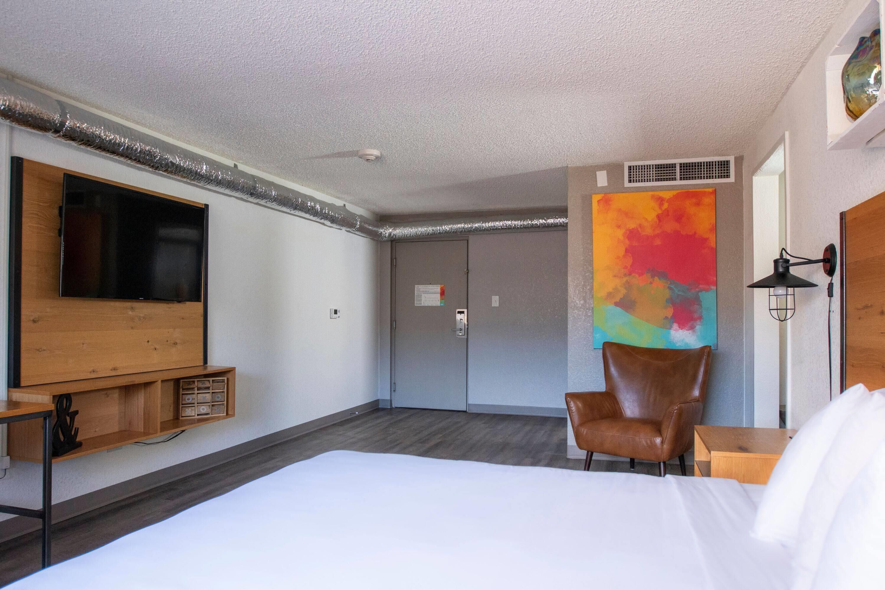 Une chambre d’hôtel avec un lit, une télévision, une chaise, un bureau et un tableau abstrait aux couleurs vives.