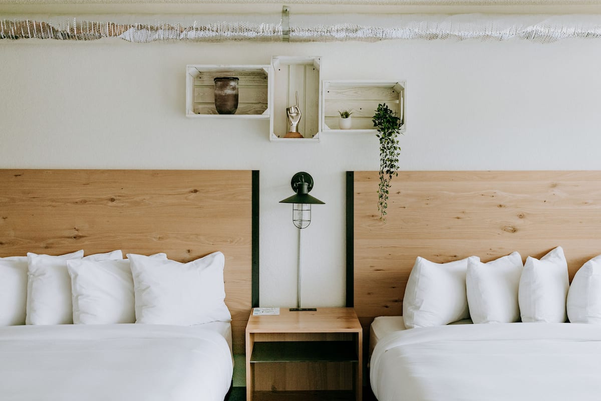 A hotel room with two beds, a nightstand, a lamp, and wall-mounted shelves with decorative items.