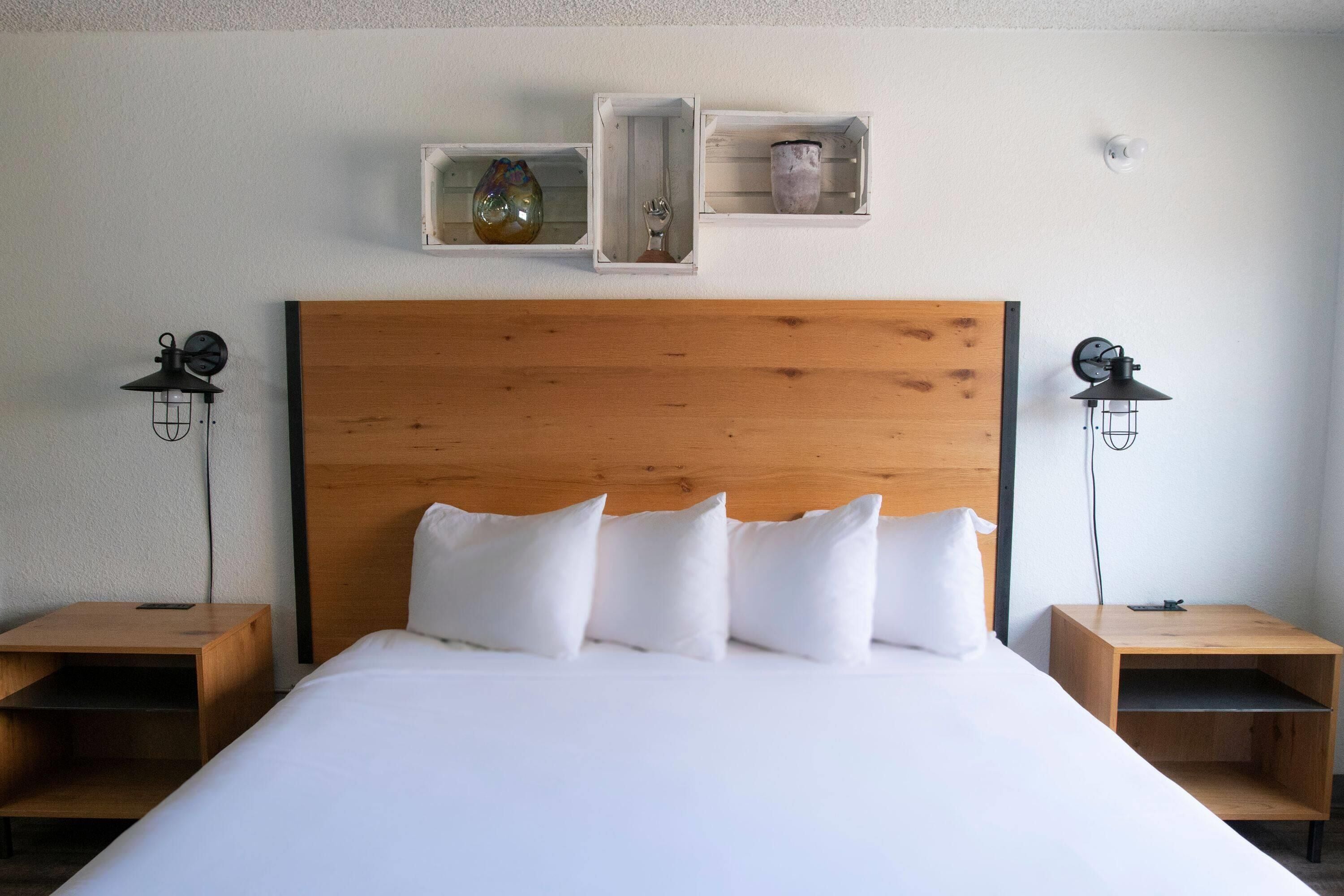Quarto de hotel com cabeceira de madeira, roupa de cama branca, duas mesas de cabeceira com abajures e prateleiras fixadas na parede com objetos decorativos.