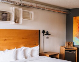 Una cama bien hecha con sábanas blancas, cabecera de madera, mesita de noche con lámpara y un pequeño mueble de madera con un cuadro abstracto de colores en la pared.