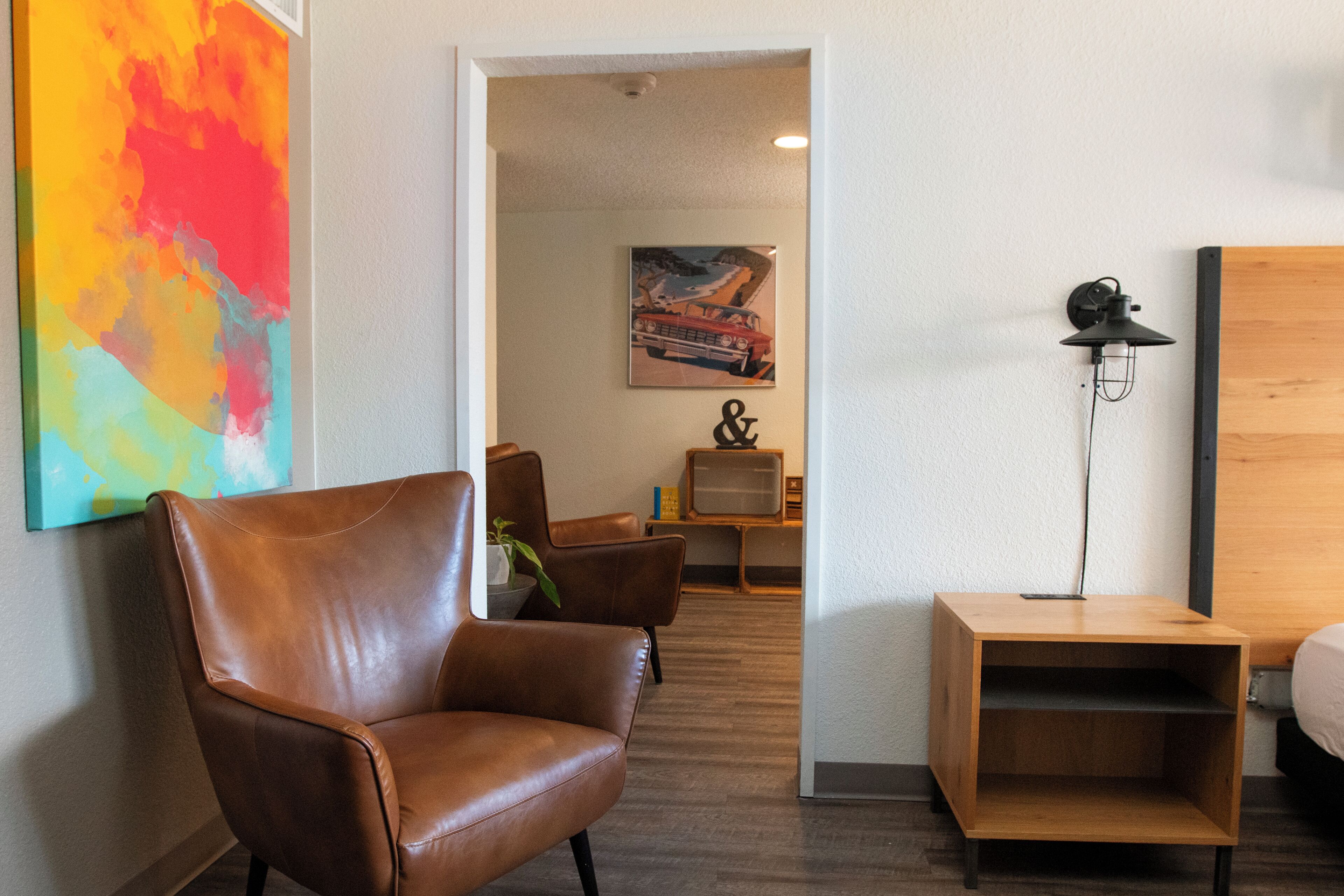 Ein Zimmer mit einem braunen Ledersessel, einem Holznachttisch, einer Stehlampe, einem Fernseher und einem farbenfrohen abstrakten Gemälde an der Wand.