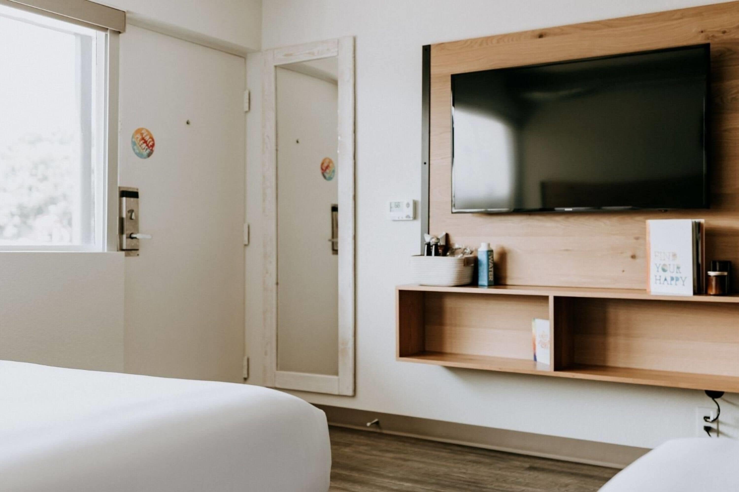 Ein Hotelzimmer mit einem großen Flachbildfernseher an einer Holzwand, einem Nachttisch mit einer Wasserflasche und einer Tür mit Griff.