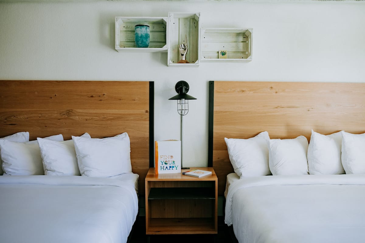 A hotel room with two beds, a nightstand with a lamp, and wall-mounted shelves with decorative items.