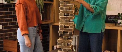 Two people playing with a tall stack of wooden blocks in a room with bookshelves and a brick wall.
