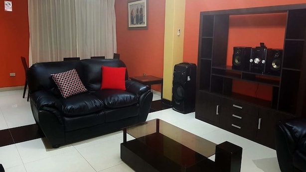 A living room with a black leather sofa, a coffee table, a TV stand with speakers, and a red accent wall. - Departamentos Perú Hosting (Piura)