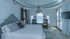 Une chambre à coucher avec un grand lit, un lustre, deux fauteuils, une télévision et une vue sur l’océan.