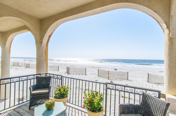 A balcony with wicker furniture and potted plants, overlooking a beach with wooden fences.
