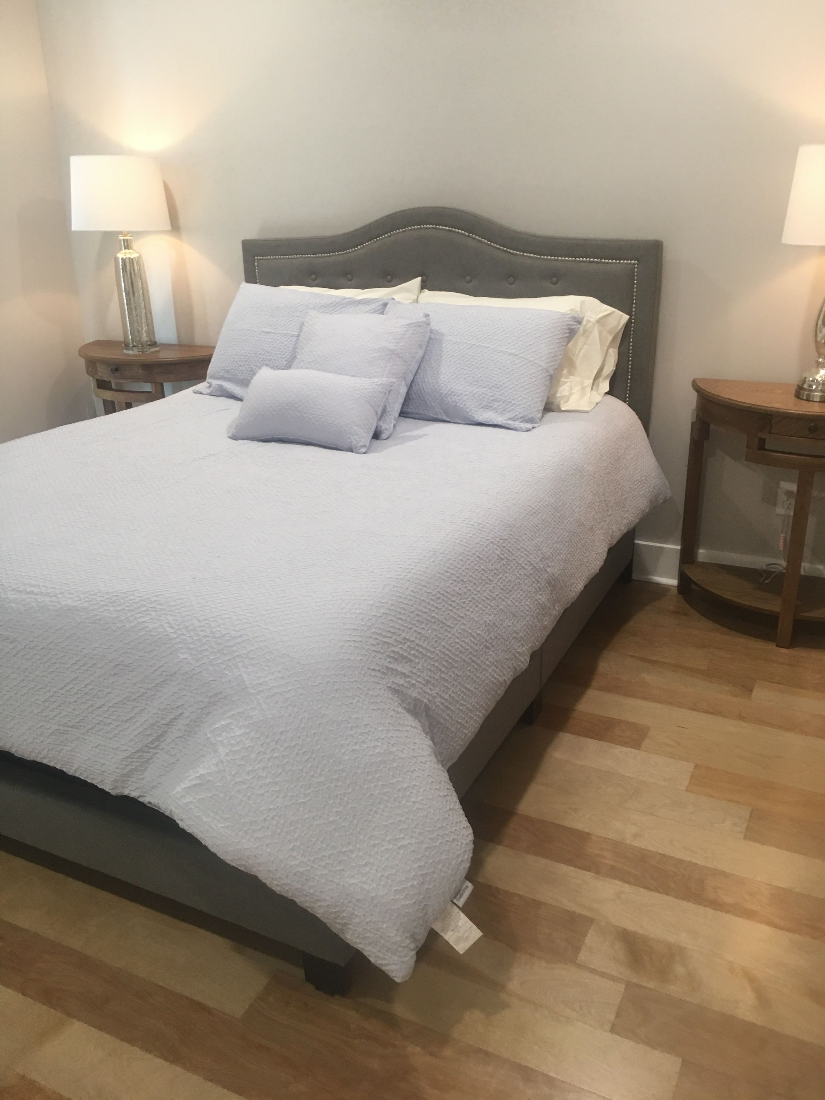 Una cama bien hecha, con cabecera gris, ropa de cama blanca y dos mesitas de noche con lámparas.