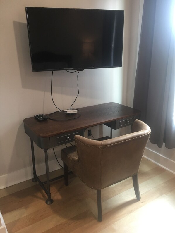 A room with a flat-screen TV mounted on the wall, a wooden desk with a chair, and a remote control on the desk.