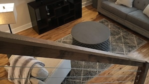 A modern living room with a flat-screen TV, a grey sofa, a patterned rug, and a staircase with a grey bench. - The Hotel at Huntington Square (Jonesboro)