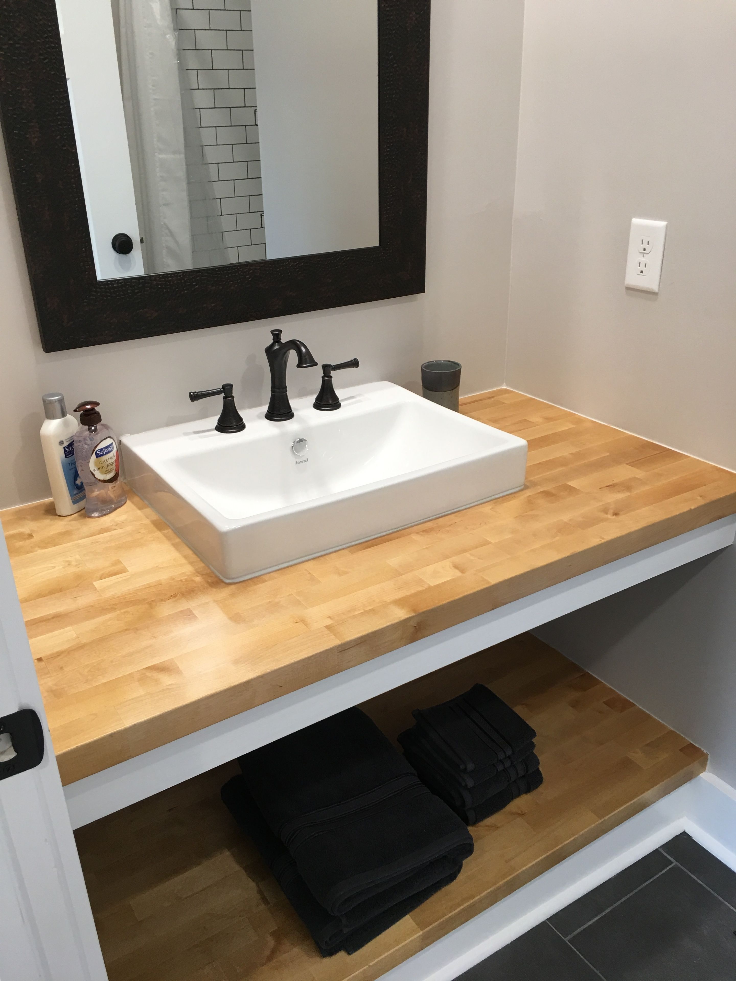 Ein Badezimmer mit einem weißen Waschbecken, schwarzen Wasserhähnen, einer Holz-Arbeitsplatte, einem Spiegel und einem Handtuchhalter mit gefalteten Handtüchern.