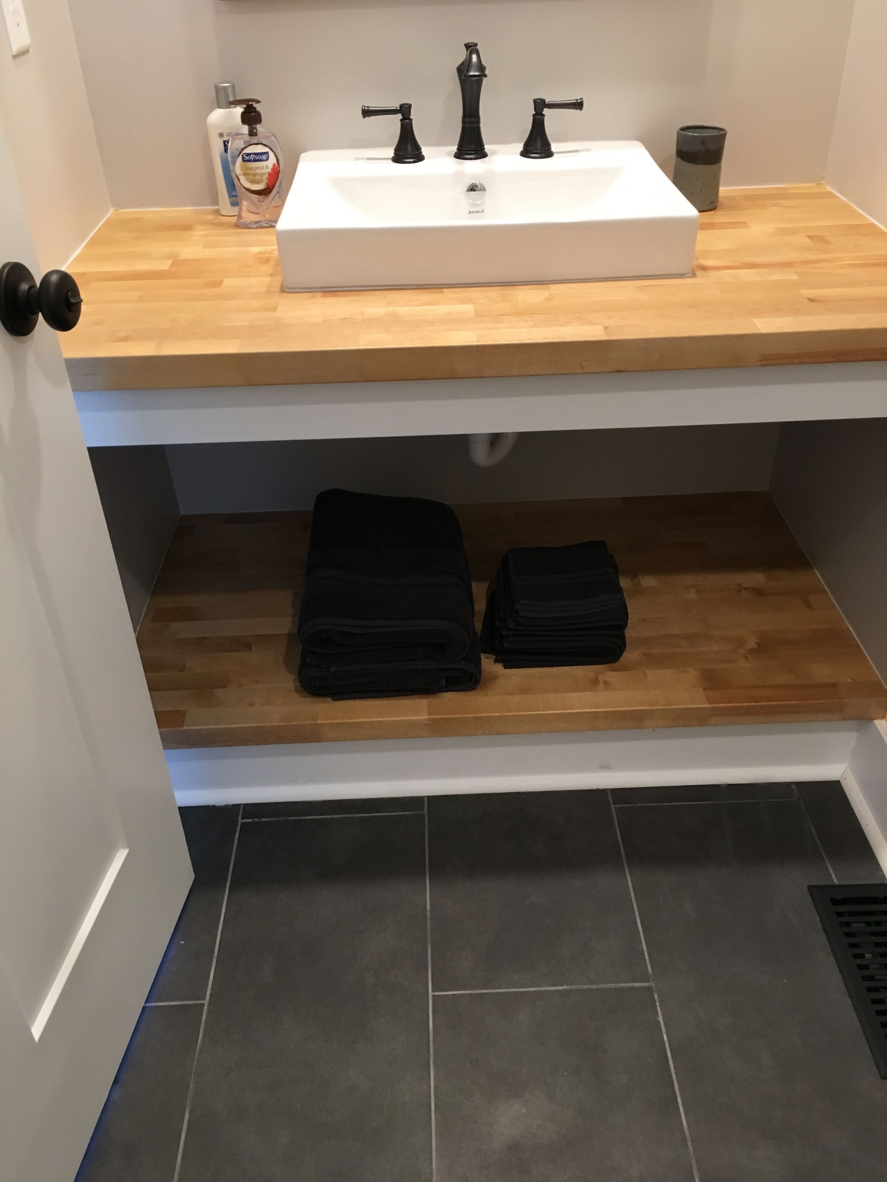A bathroom with a white sink, wooden countertop, and two black towels on a shelf.