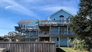 A two-story building with a balcony on the second floor, a wooden deck in front, and a clear blue sky. - 3rd row Whalehead house w/ private pool, hot tub, elevator, pool table, pet friendly (Corolla)
