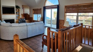 A spacious living room with a large sectional sofa, a wooden staircase, and a balcony with outdoor seating. - 3rd row Whalehead house w/ private pool, hot tub, elevator, pool table, pet friendly (Corolla)