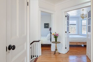 Un pasillo que conduce a un dormitorio con piso de madera, una pequeña mesa redonda con un florero y una cama con edredón blanco.