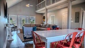 A spacious living area with a high ceiling, a dining table with red chairs, a sofa with blue and gray cushions, and a staircase leading to an upper level. - Spacious, budget friendly 3 BR House! (Gulf Shores)