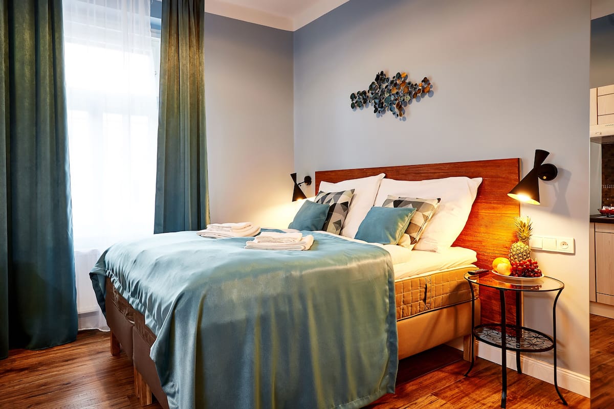 A neatly made bed with a blue bedspread, a wooden headboard, and a small table with fruit and a remote control beside it.