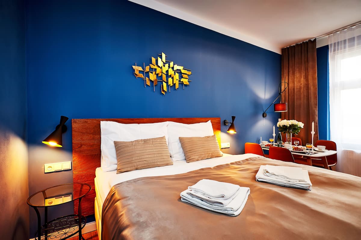 A hotel room with a large bed, a wall art piece, a small table with a lamp, and a dining area with a table set for two.