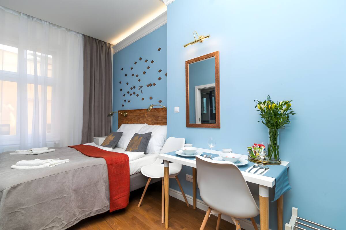 A hotel room with a bed, a dining table with chairs, a mirror, and a vase with flowers.