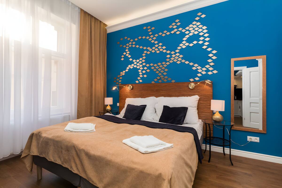 A hotel room with a bed, two bedside tables, a wall art piece, a mirror, and a window with curtains.