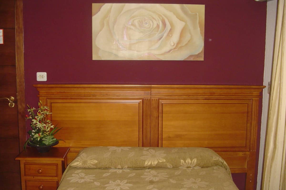 A bedroom with a wooden headboard, a floral bedspread, a nightstand with a plant, and a large rose painting on the wall.