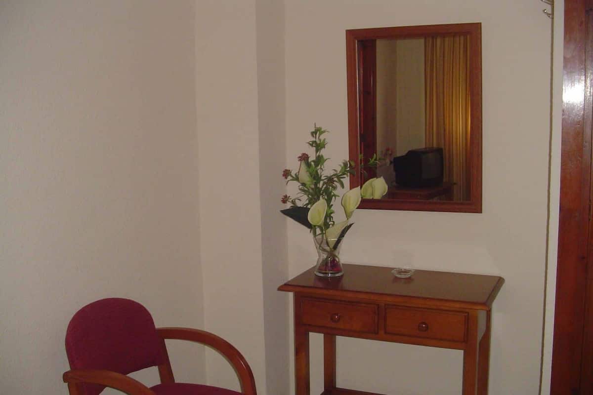 A small room with a wooden desk, a chair, a mirror, and a vase with flowers.