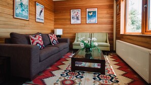 Un salon chaleureux avec un canapé marron, un fauteuil vert et une table basse en verre. Des affiches ornent les murs en bois et un tapis à motifs recouvre le sol.