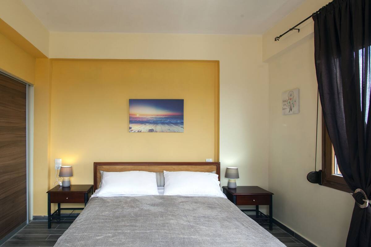 A hotel room with a bed, two bedside tables with lamps, a painting on the wall, and a window with curtains.