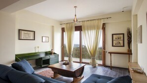 A living room with a sofa, coffee table, and a view of the outdoors through a window with curtains. - Foivos and Athina Villas (Archanes-Asterousia)