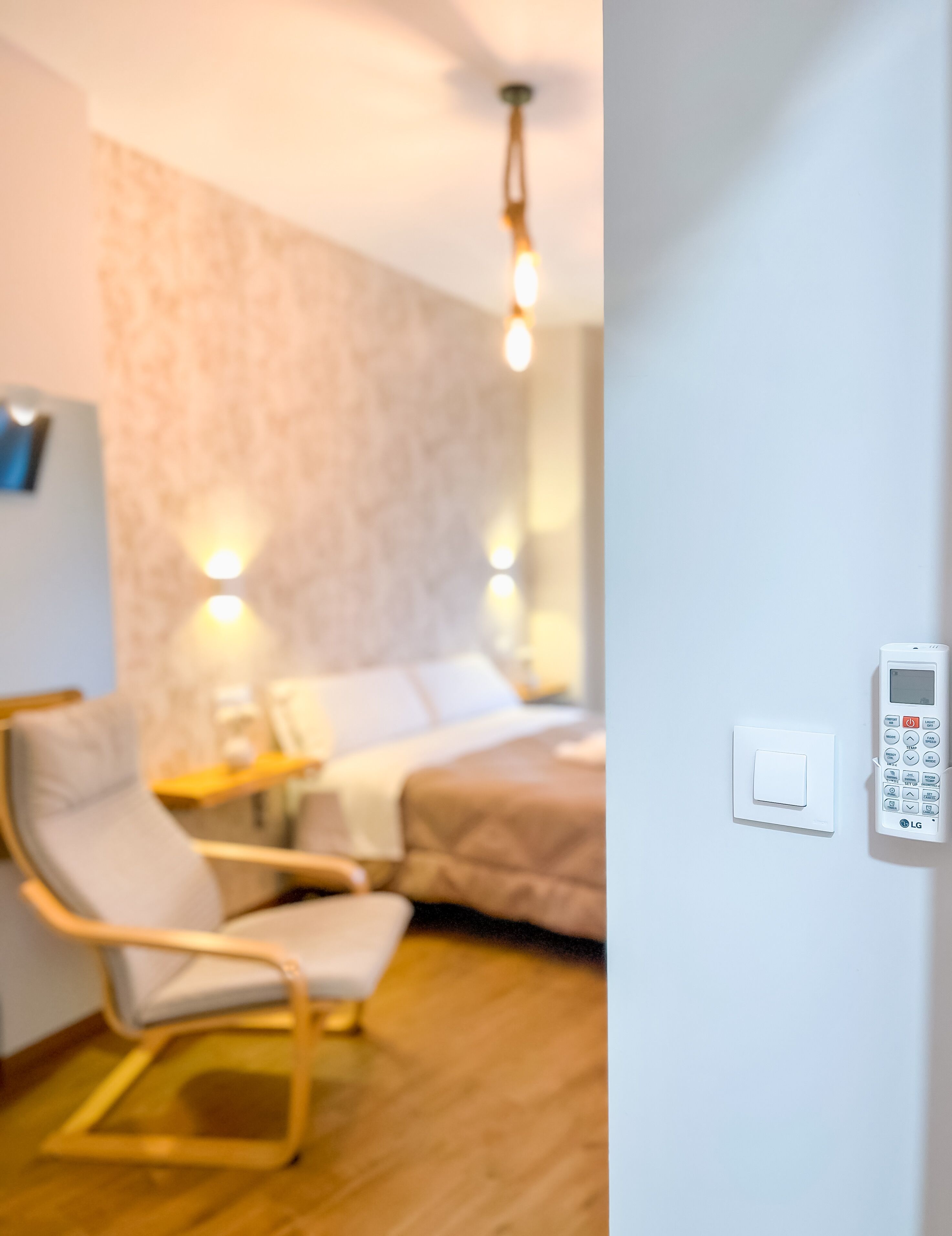 A modern hotel room with a bed, a chair, a bedside table, and a wall-mounted control panel.