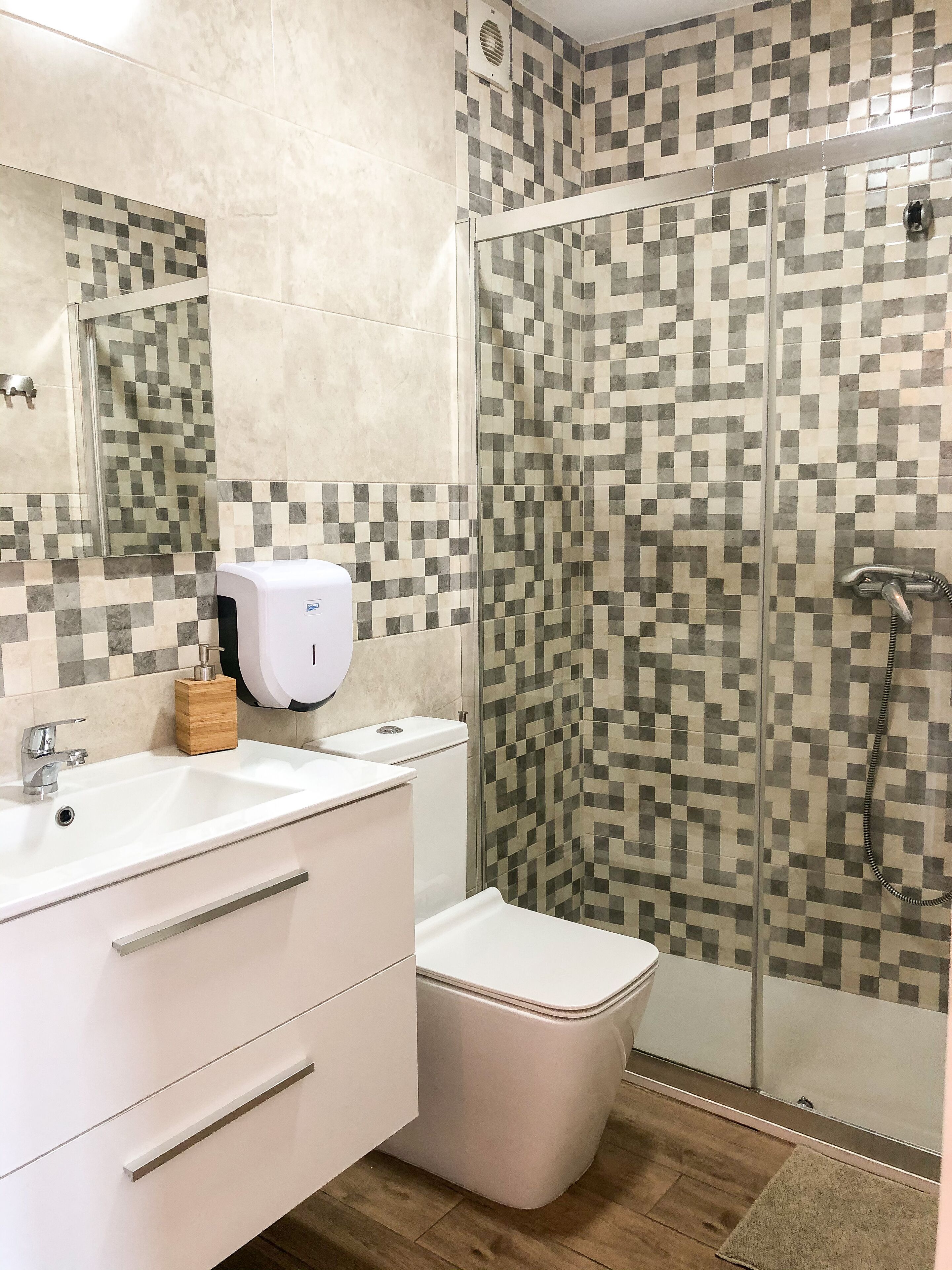 Une salle de bain avec un lavabo blanc, des toilettes et une douche dont le mur est recouvert de carreaux à motifs géométriques.