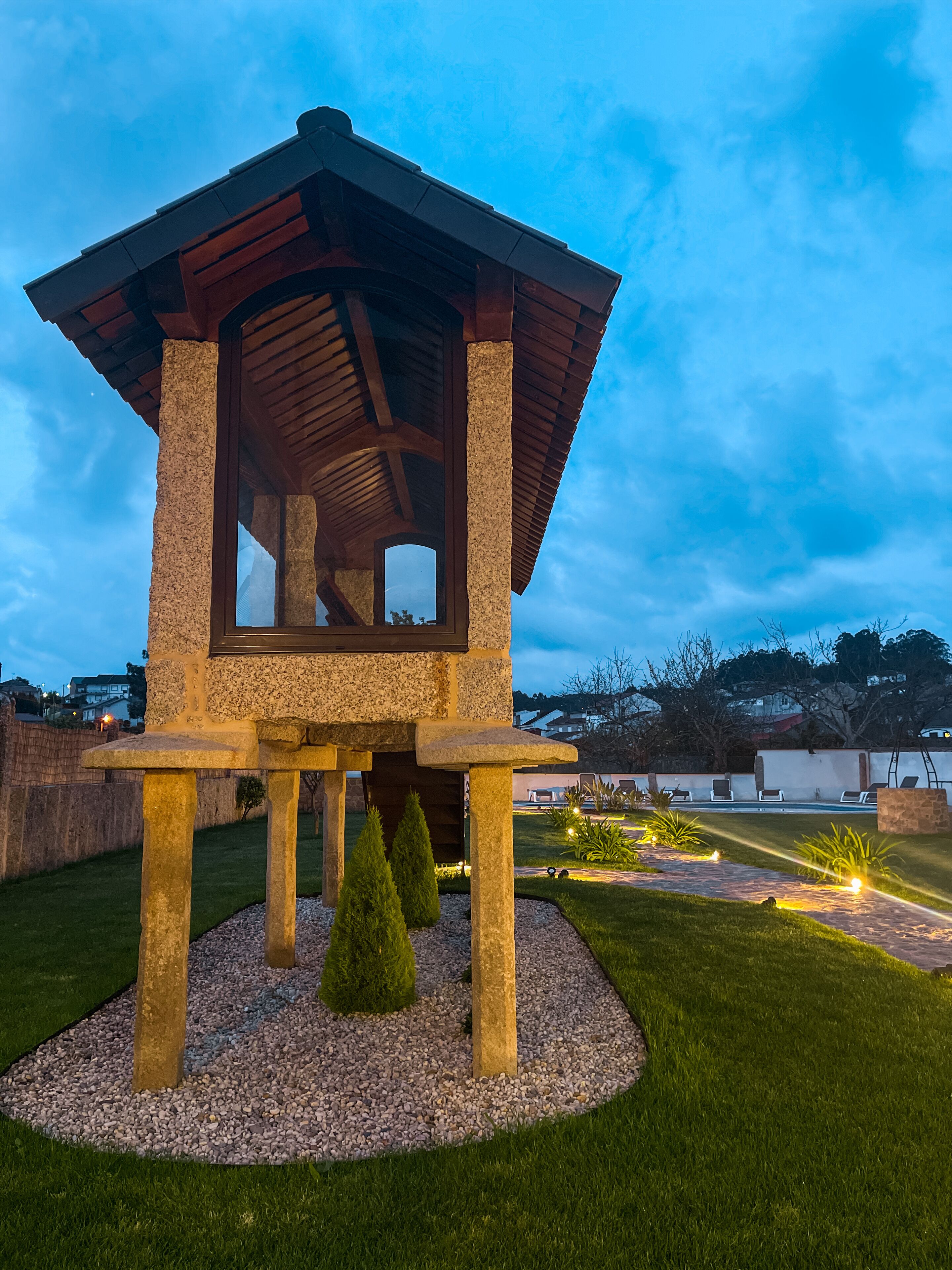 A stone gazebo with a wooden roof, surrounded by a gravel area and small trees, set in a landscaped garden with a pool and lounge chairs in the background.