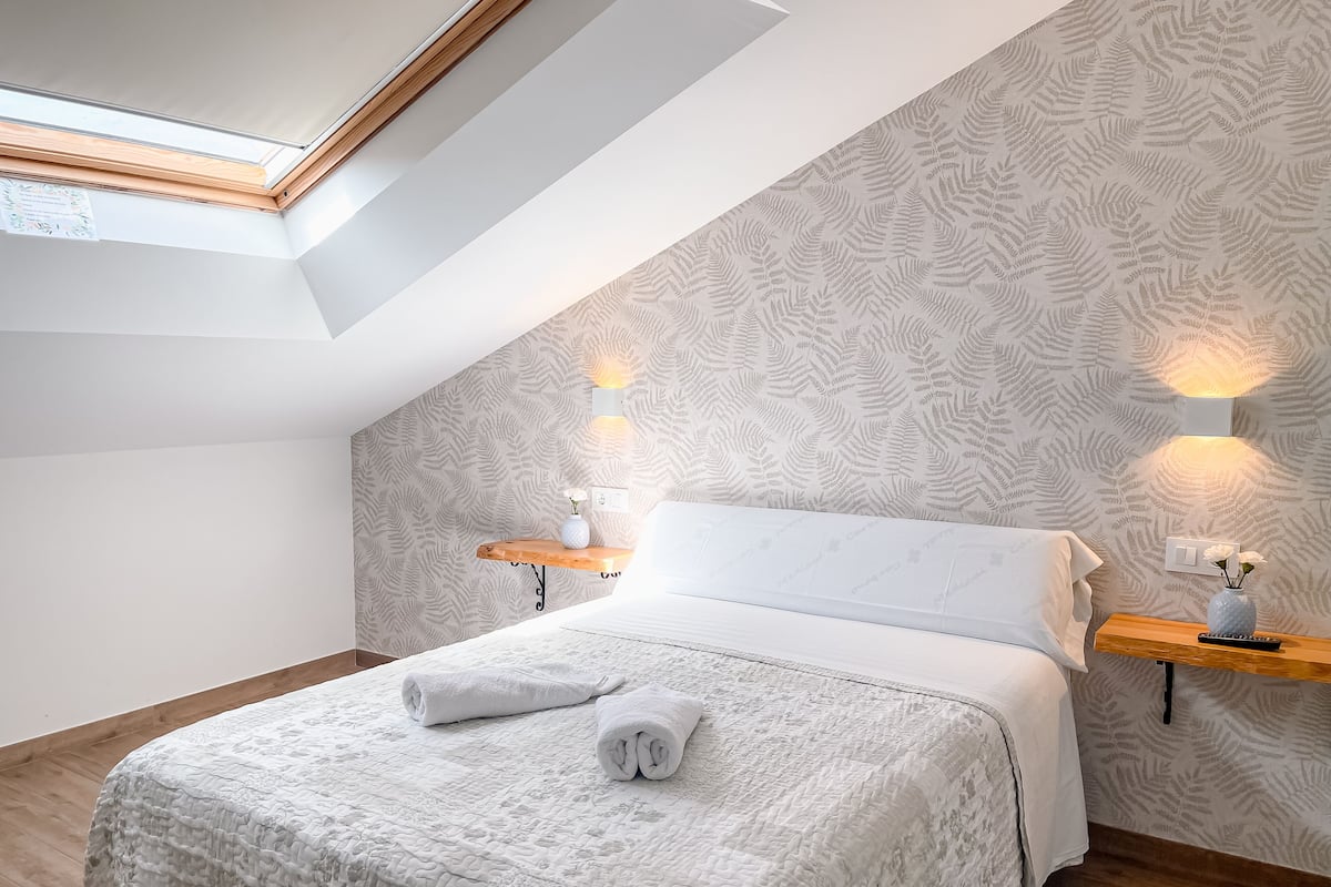 Une chambre avec un plafond en pente, un puits de lumière, un lit recouvert d’une courtepointe, deux tables de chevet avec des lampes et un papier peint à motifs.