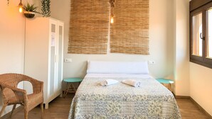 A room with a bed, a wicker chair, a small table, and a wall-mounted light fixture.