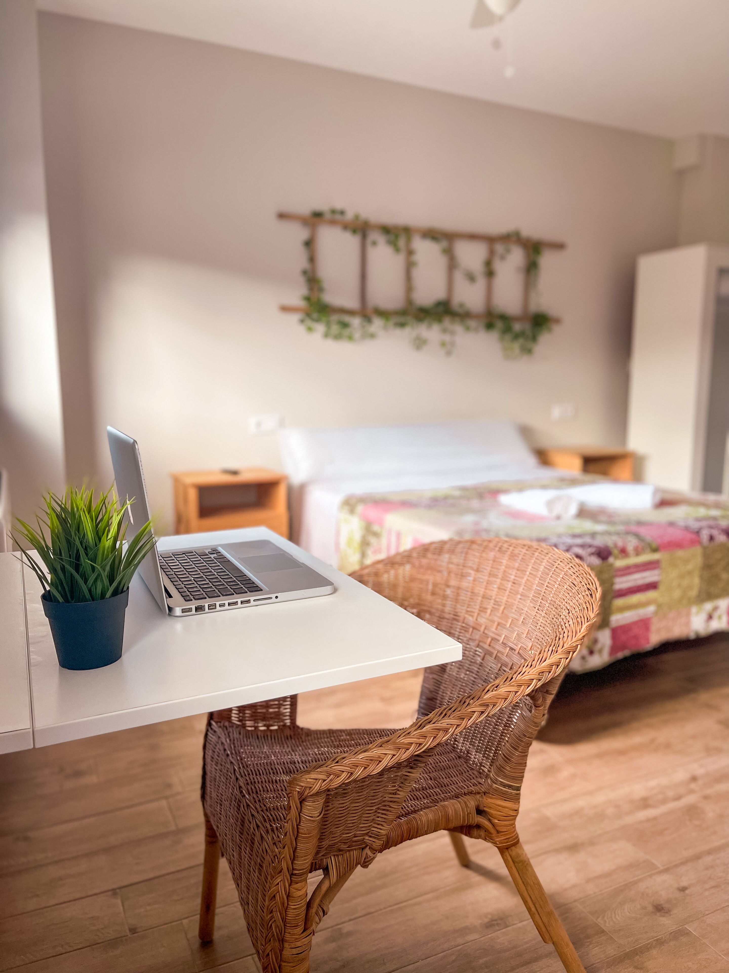 A room with a laptop on a white table, a wicker chair, a bed with a colorful quilt, and a wall-mounted plant.