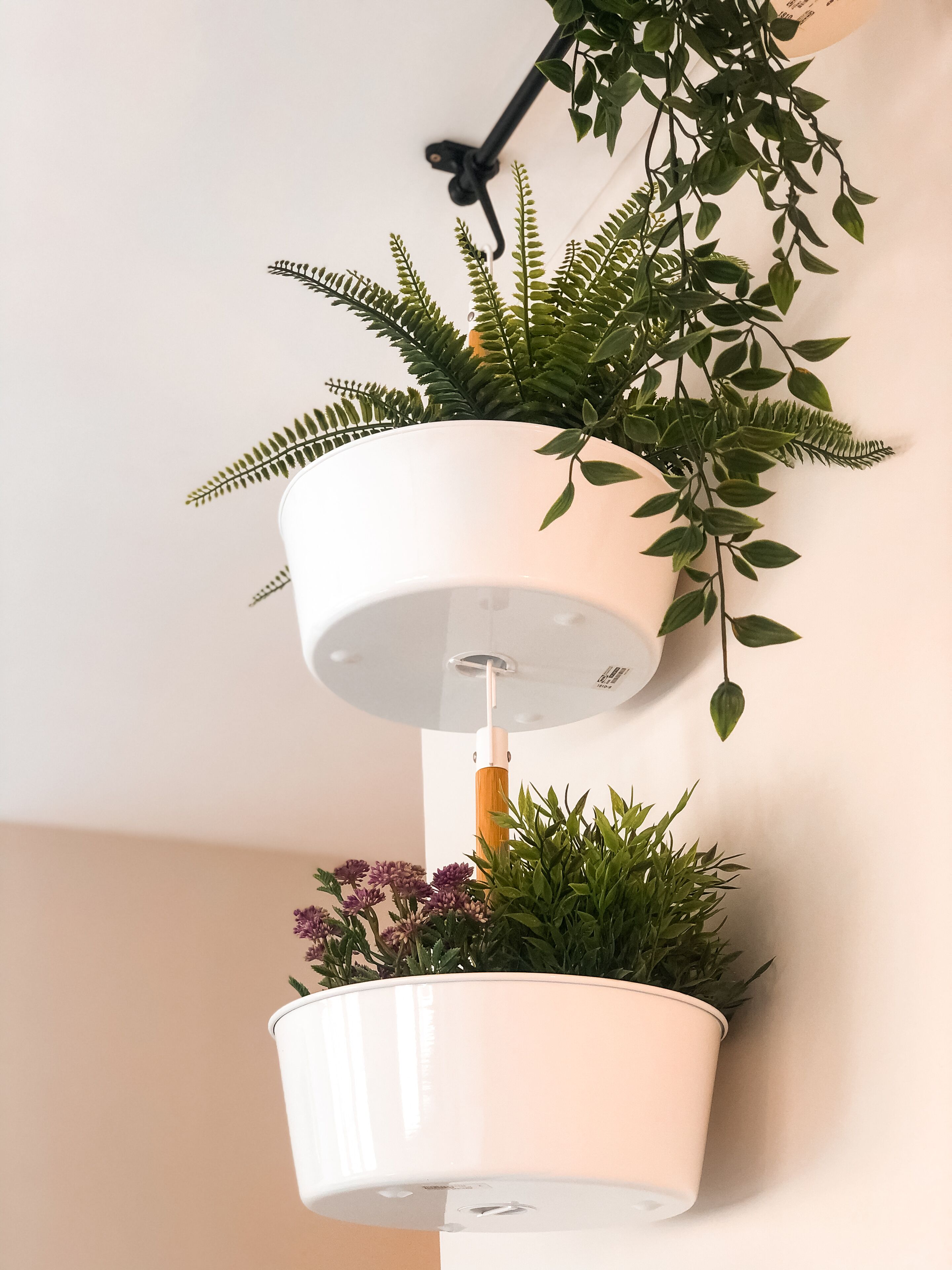 Pared branca com dois vasos suspensos brancos, um acima do outro, cada um contendo plantas verdes e flores roxas.