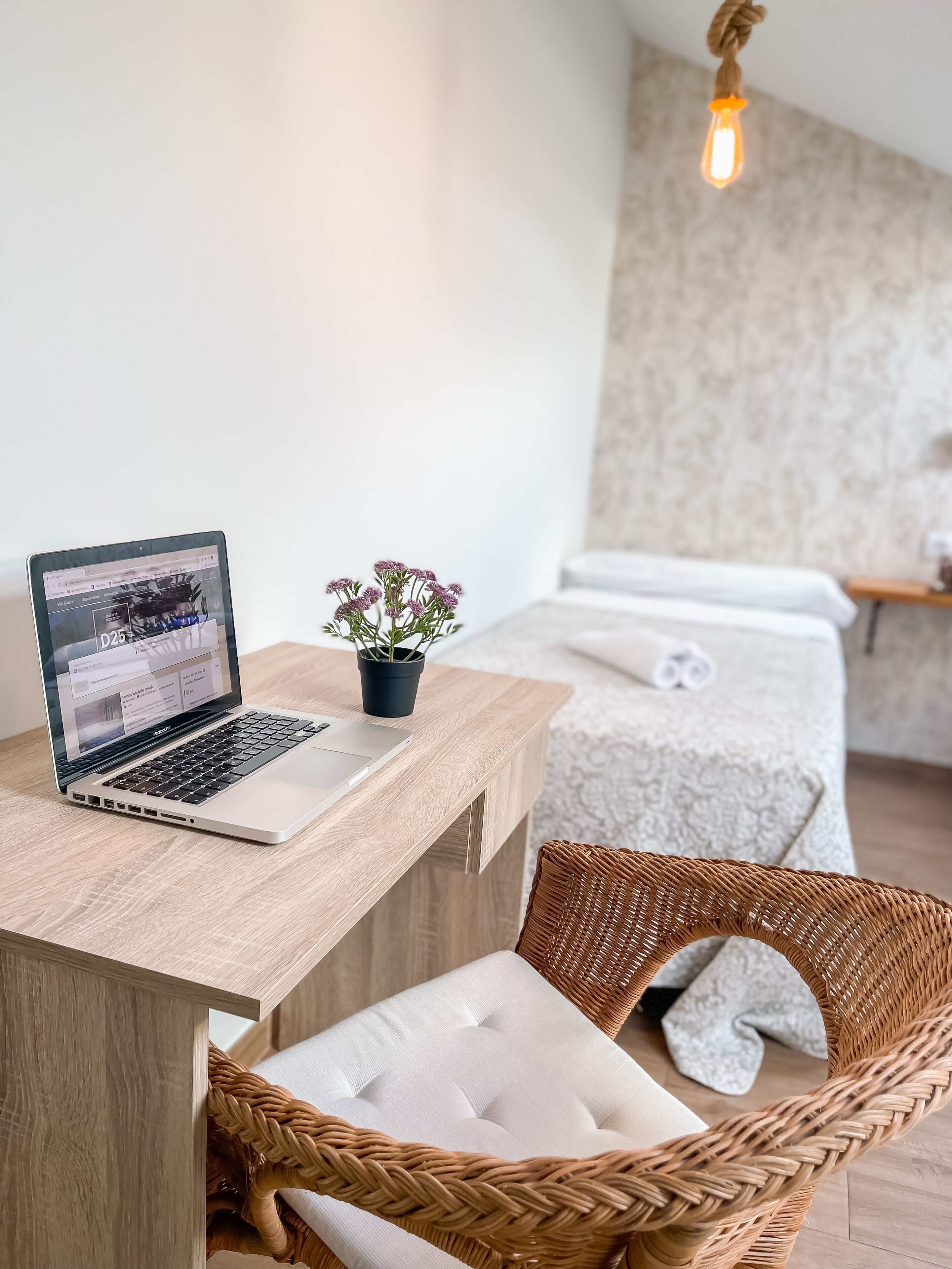 Una computadora portátil sobre un escritorio de madera, una planta pequeña en maceta, una silla de mimbre y una mesa de masajes con una toalla.